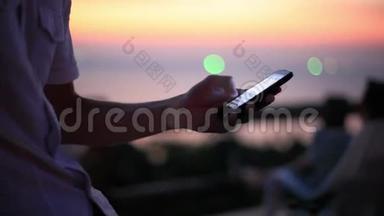 人类`手在日落海滩海边使用移动智能手机。 有模糊的bokeh灯。 1920x1080
